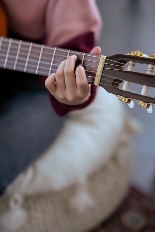 Free Unrecognizable female musician with fingers on chord playing acoustic guitar while sitting on comfortable pouf on blurred background at home Stock Photo