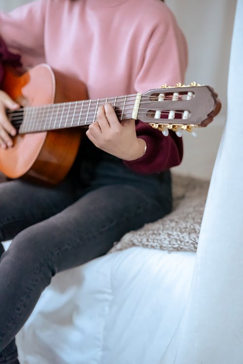 Unrecognizable female musician playing acoustic guitar while sitting on comfortable bed in cozy bedroom with white curtains during rehearsal at home