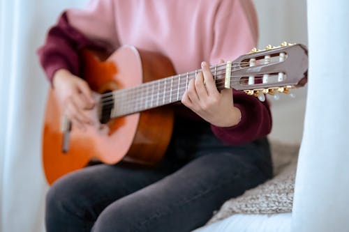 Faceless skilled female musician playing acoustic guitar while sitting on comfortable bed near white curtains during music performance at home