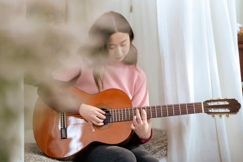 Talented Asian female musician playing acoustic guitar while sitting on soft bed in bedroom with curtains and bouquet of flowers