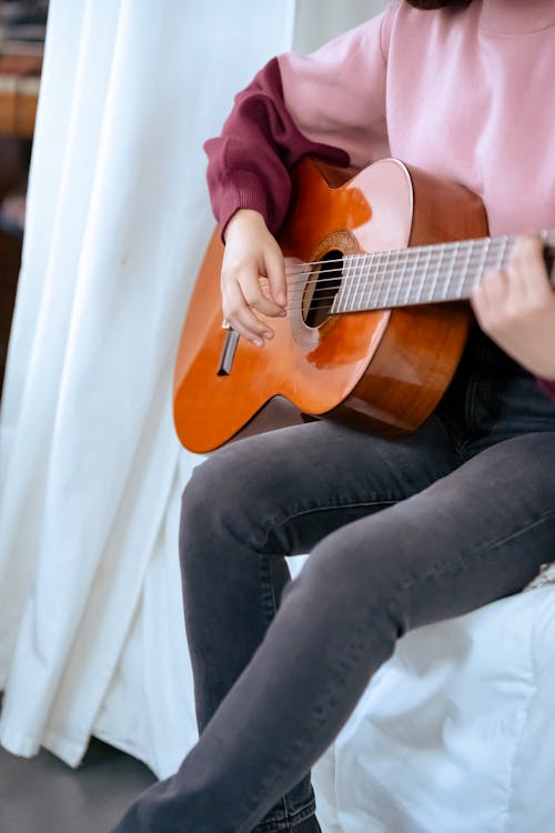 Unrecognizable female guitarist playing acoustic instrument while sitting on comfortable bed near white curtains in cozy bedroom during rehearsal at home