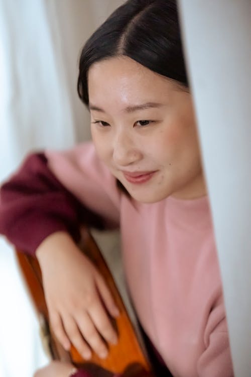 From above of positive Asian female musician playing acoustic guitar while sitting near white curtains on blurred background at home