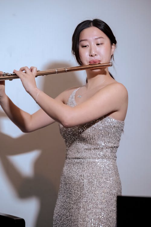 Elegant Asian female musician in elegant dress standing on stage at spotlight and playing flute during performance