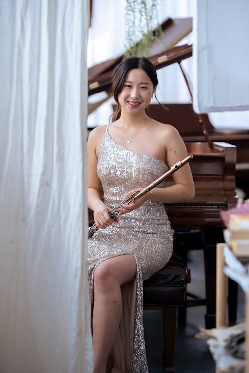 Young Asian female musician in elegant dress looking at camera while sitting near piano and holding flute before concert