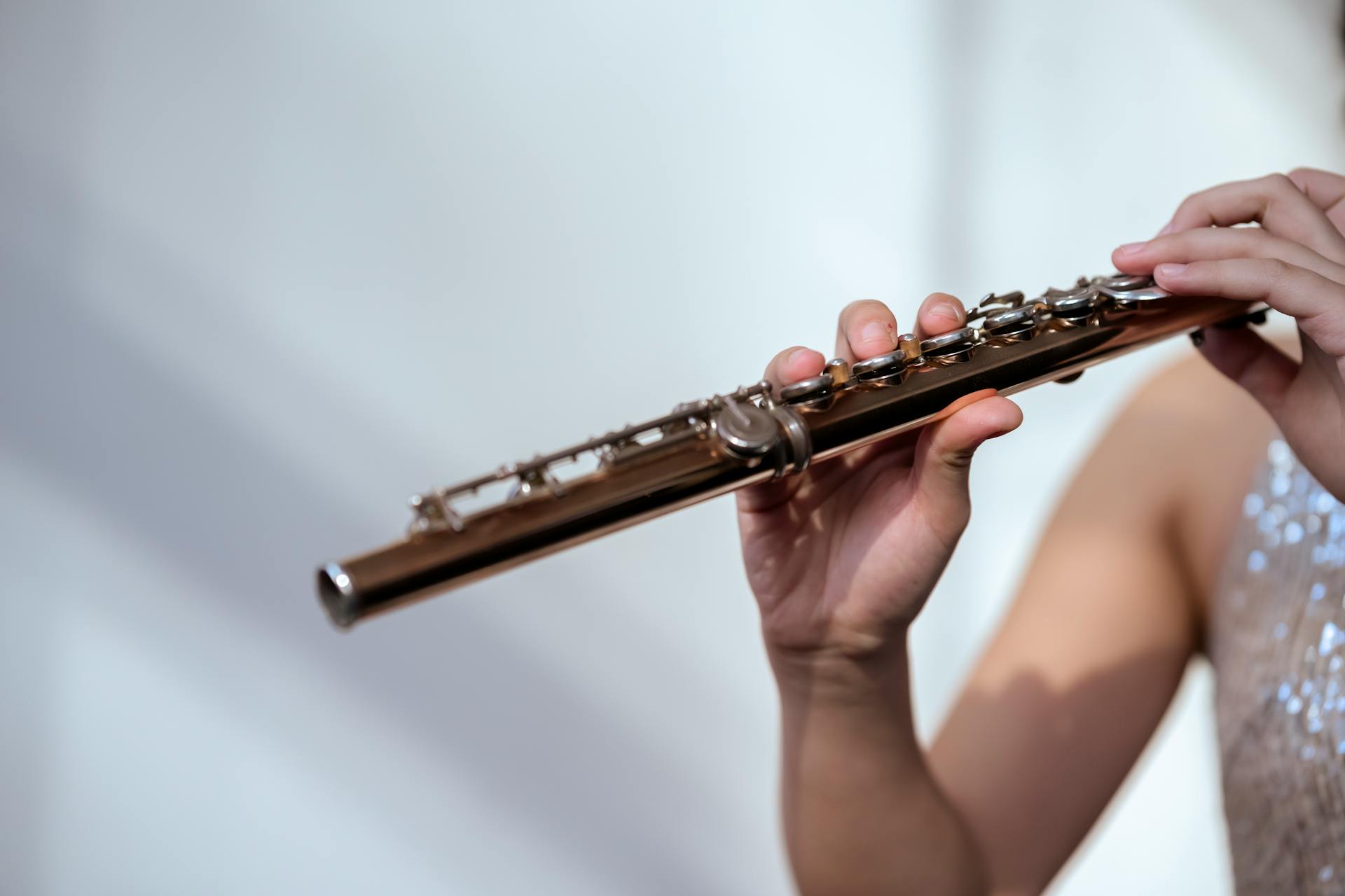 Crop unrecognizable female musician in glittering dress with bare shoulder playing flute on white background