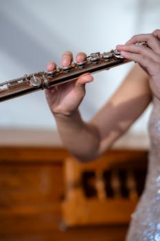 Young female musician in shiny dress playing flute near piano in light room during rehearsal for concert