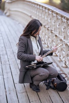 Young Asian female musician in gray suit and white blouse squatting on shabby plank footbridge and getting flute out of instrument case