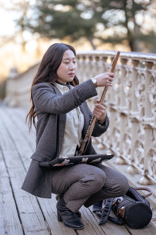 Focused Asian female with long loose hair squatting on wooden bridge near railing in park and assembling flute