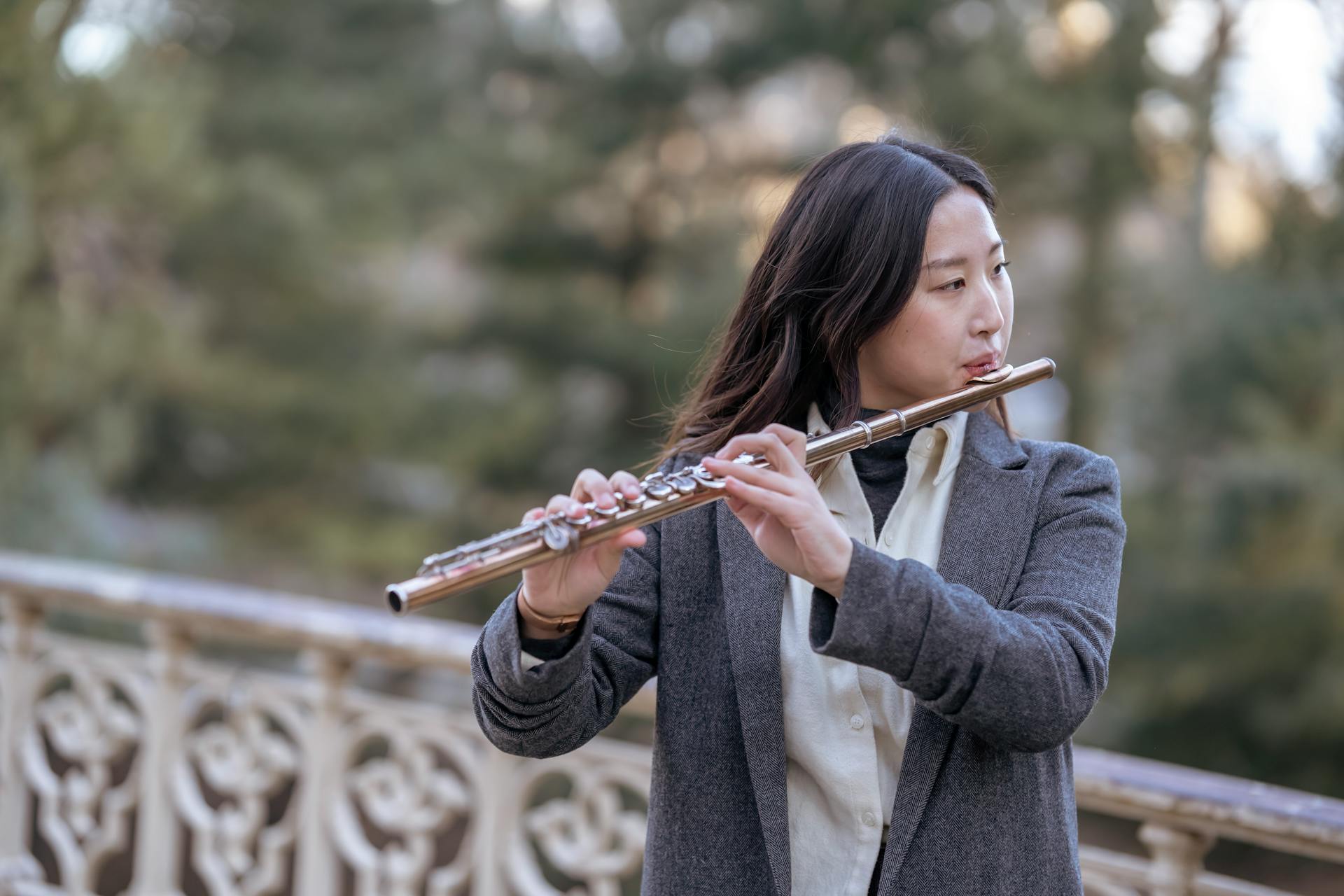 A young woman plays the flute outdoors in a serene setting, wearing a blazer.