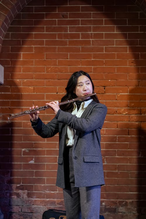 Elegant Asian female musician standing on street against brick wall and playing flute