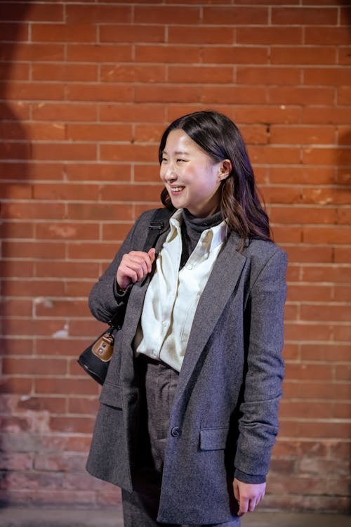 Positive young ethnic female wearing classy outfit standing with bag against brick wall and looking away