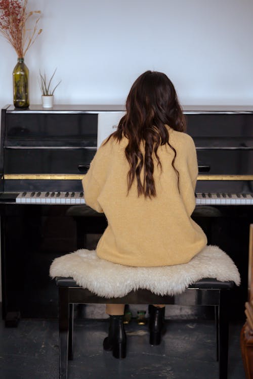 Back view full body of unrecognizable woman with long hair sitting on stool and playing piano