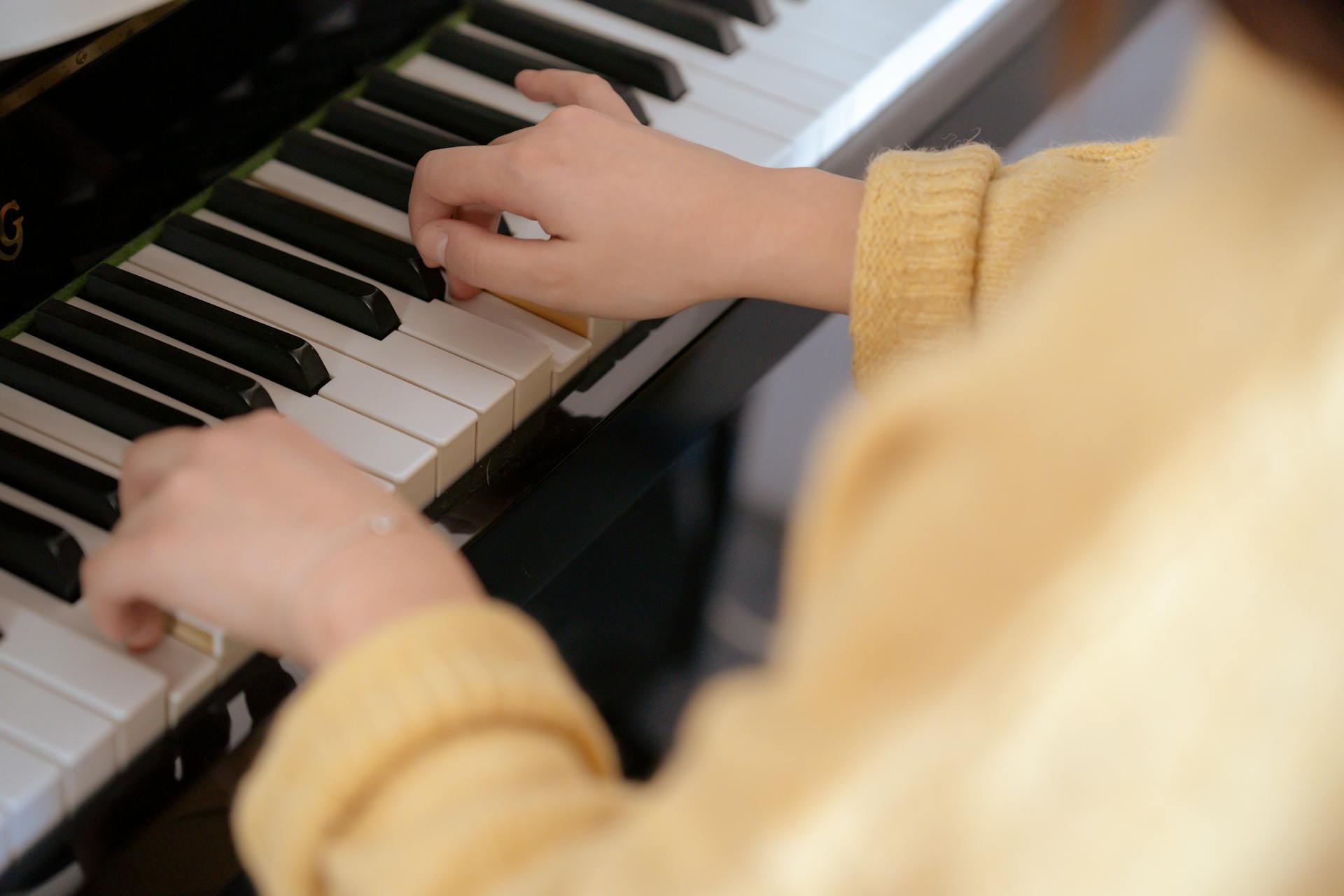 From above of crop unrecognizable female musician in warm pullover sitting on piano stool and touching black and white keys