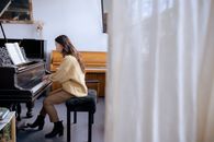 Full body of concentrated Asian lady in casual clothes and boots on high heels sitting on stool and gently touching piano keys and attentively looking at music book