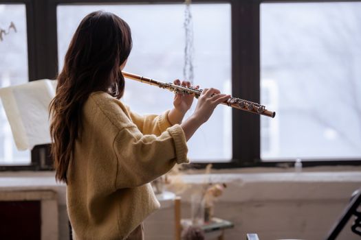Side view of anonymous lady in casual sweater playing flute while standing near windows in light room