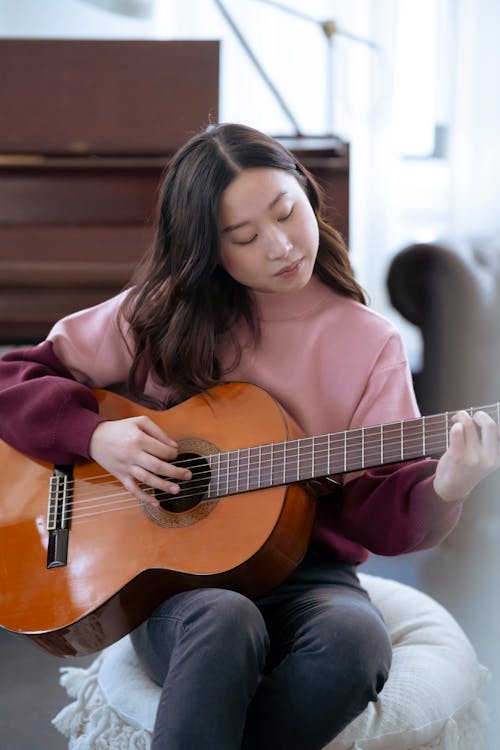 Free Ethnic female playing guitar on pouf in room Stock Photo