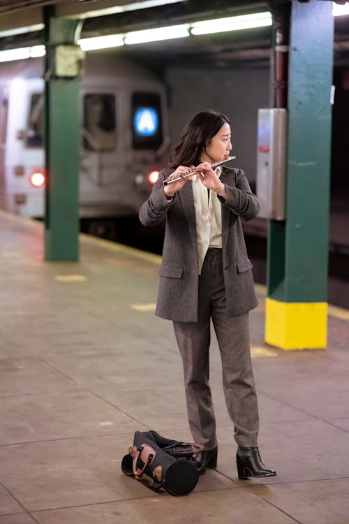 Full body of ethnic young woman in elegant suit playing flute on subway station near column and train
