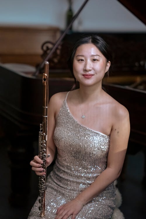 Positive young Asian lady in elegant dress sitting with flute in hand looking at camera in light room near piano