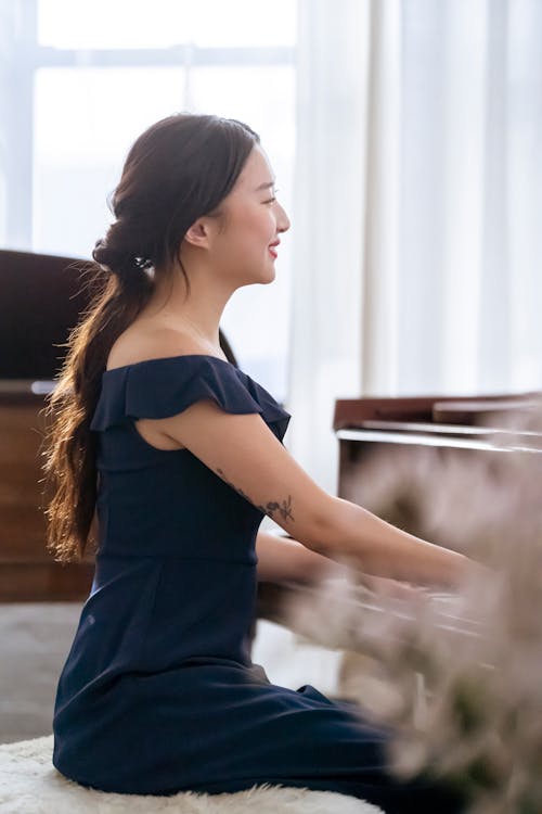 Free Side view of positive young woman in elegant dress playing piano while sitting on pouf in bright studio near windows with curtains Stock Photo