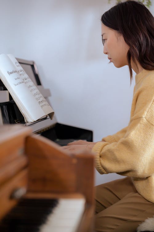 Free Side view of concentrated young Asian lady in casual clothes playing piano while sitting in bright studio with music book Stock Photo