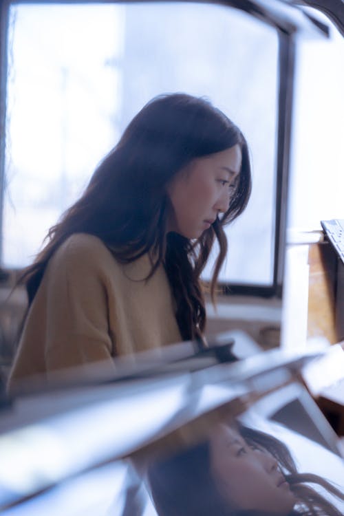 Reflection of concentrated young Asian lady in casual clothes playing piano in bright studio
