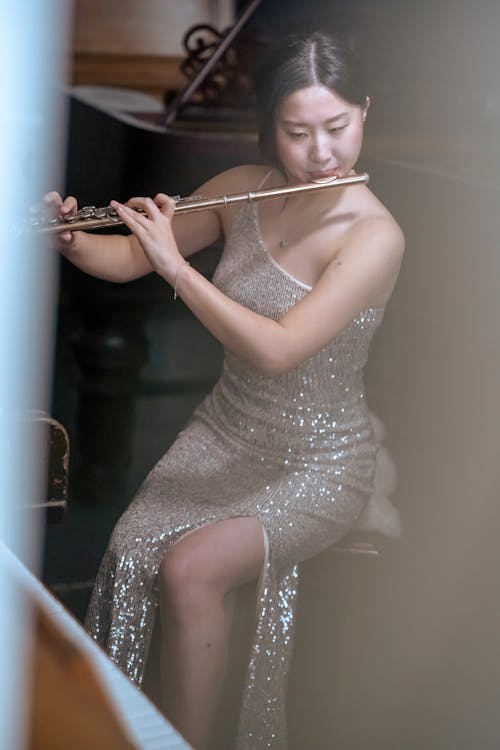 Young Asian female flutist in shiny dress playing flute and looking away while preparing for concert