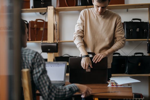 Young artisan selecting leather briefcase in workshop with laptop and numerous bags displayed in the background.