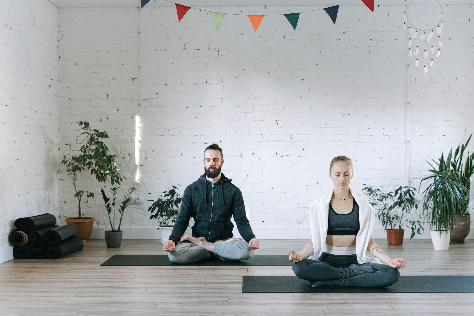 Two adults meditating in lotus pose indoors, surrounded by plants, promoting serenity and wellness.