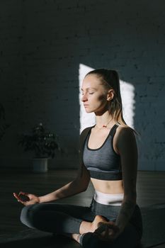 A serene young woman meditates indoors with eyes closed, basking in natural light.