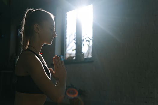 A young woman practicing meditation indoors with sunlight streaming in through a window.