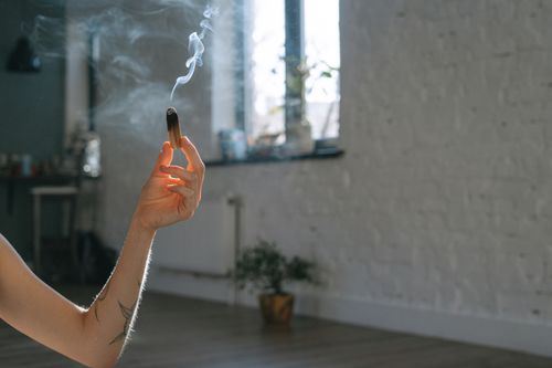 Free A man's hand holding a burning Palo Santo stick with smoke wafting in a sunlit room, creating a serene atmosphere. Stock Photo