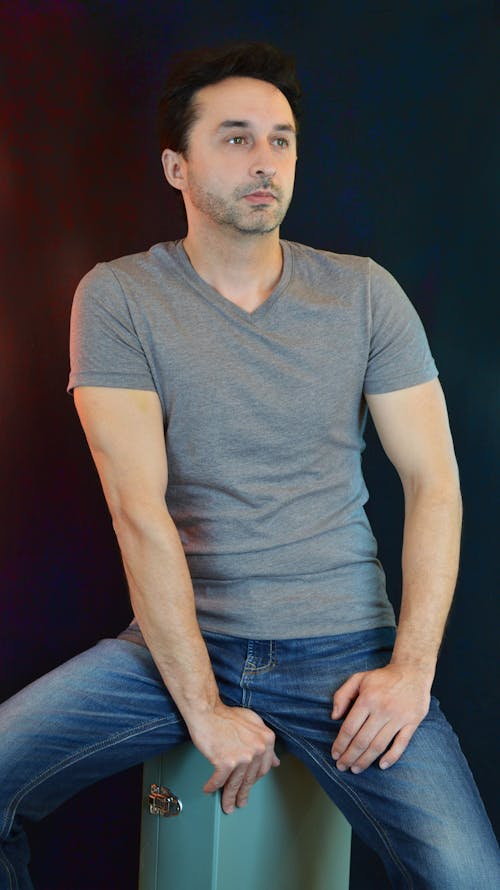 Self assured young male model in casual outfit sitting on stool and looking away thoughtfully against black background