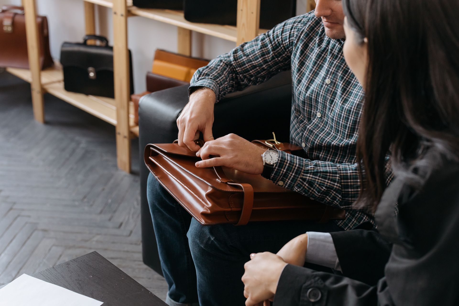 A man demonstrates a brown leather briefcase to a woman in a stylish showroom.