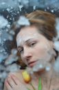Top view of tranquil frozen female touching bare shoulders while lying under ice with fresh tulip in hand and looking at camera