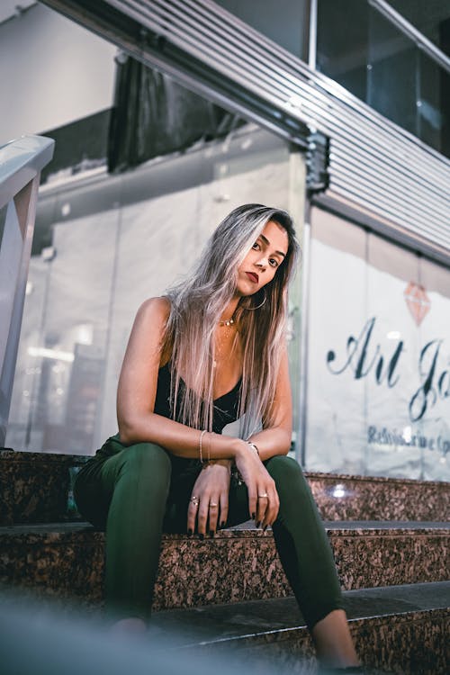 Young female with long dyed hair looking at camera and resting on stairs of contemporary building