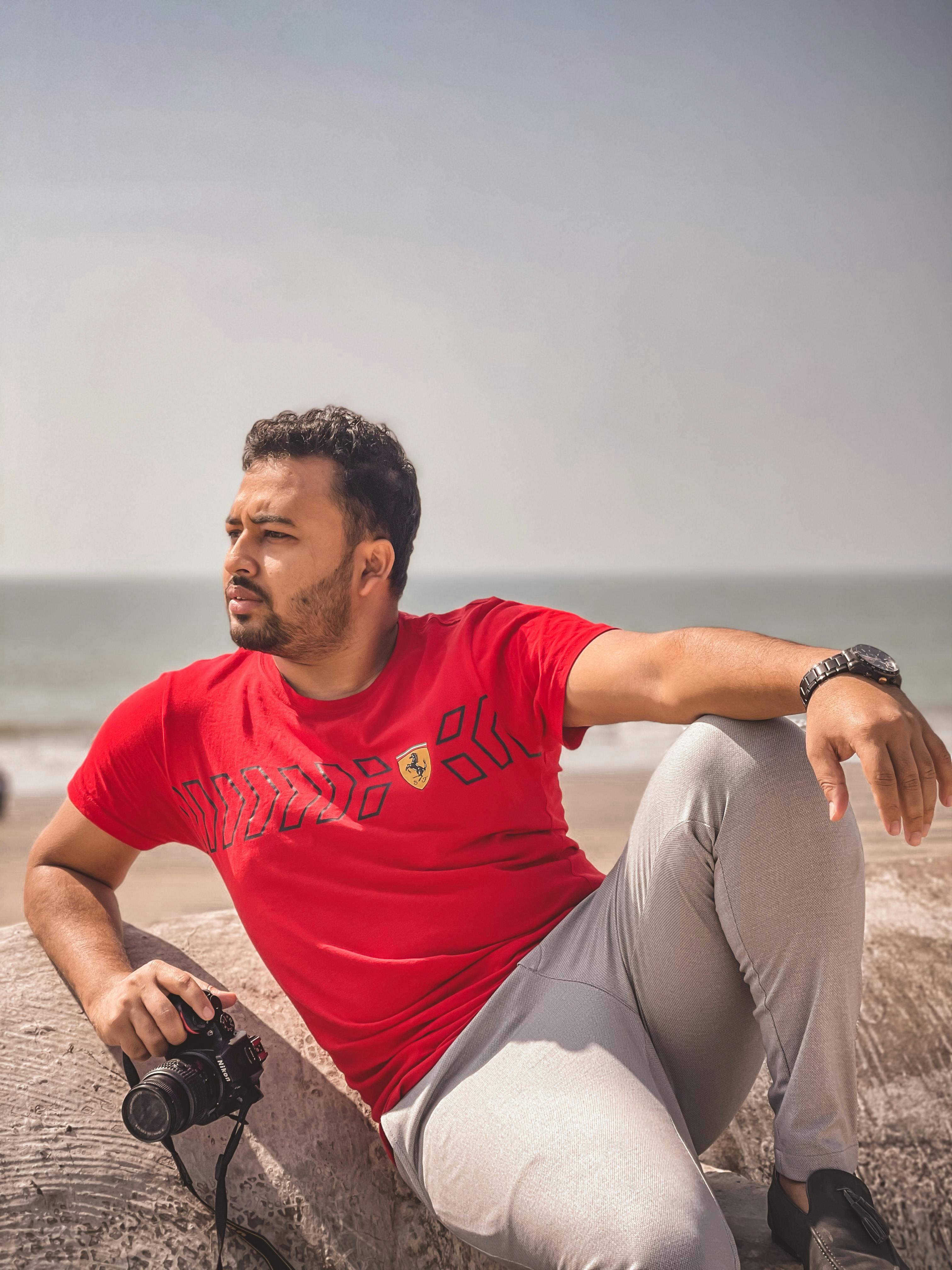 Man in red t-shirt sitting on beach, holding a camera. Warm and relaxed setting.