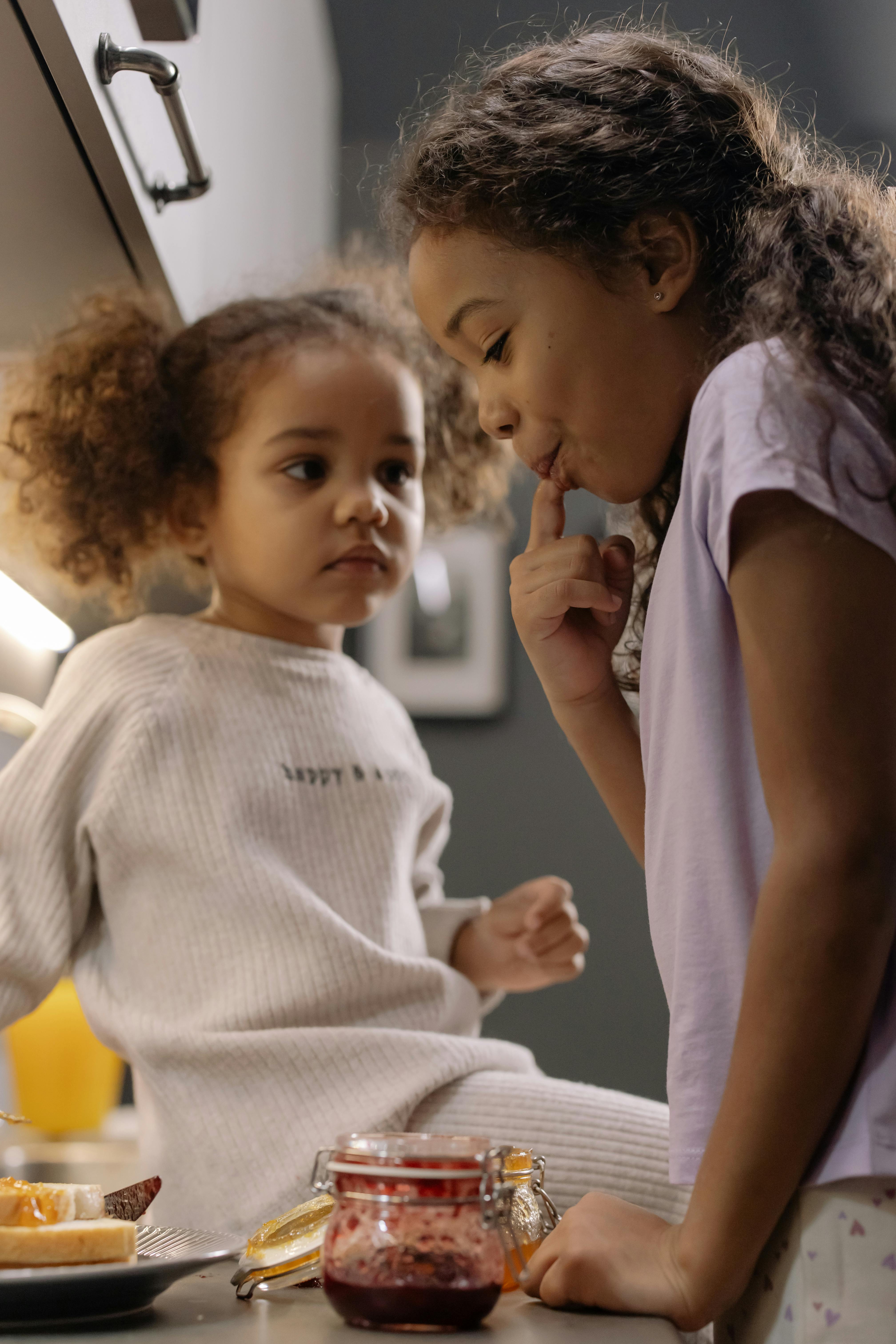 Two young sisters enjoy making and tasting jam sandwiches in a cozy kitchen setting.