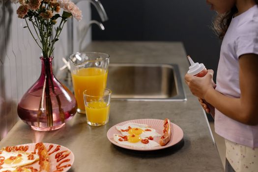 A child decorates breakfast with ketchup by a kitchen sink, with juice and fresh flowers.