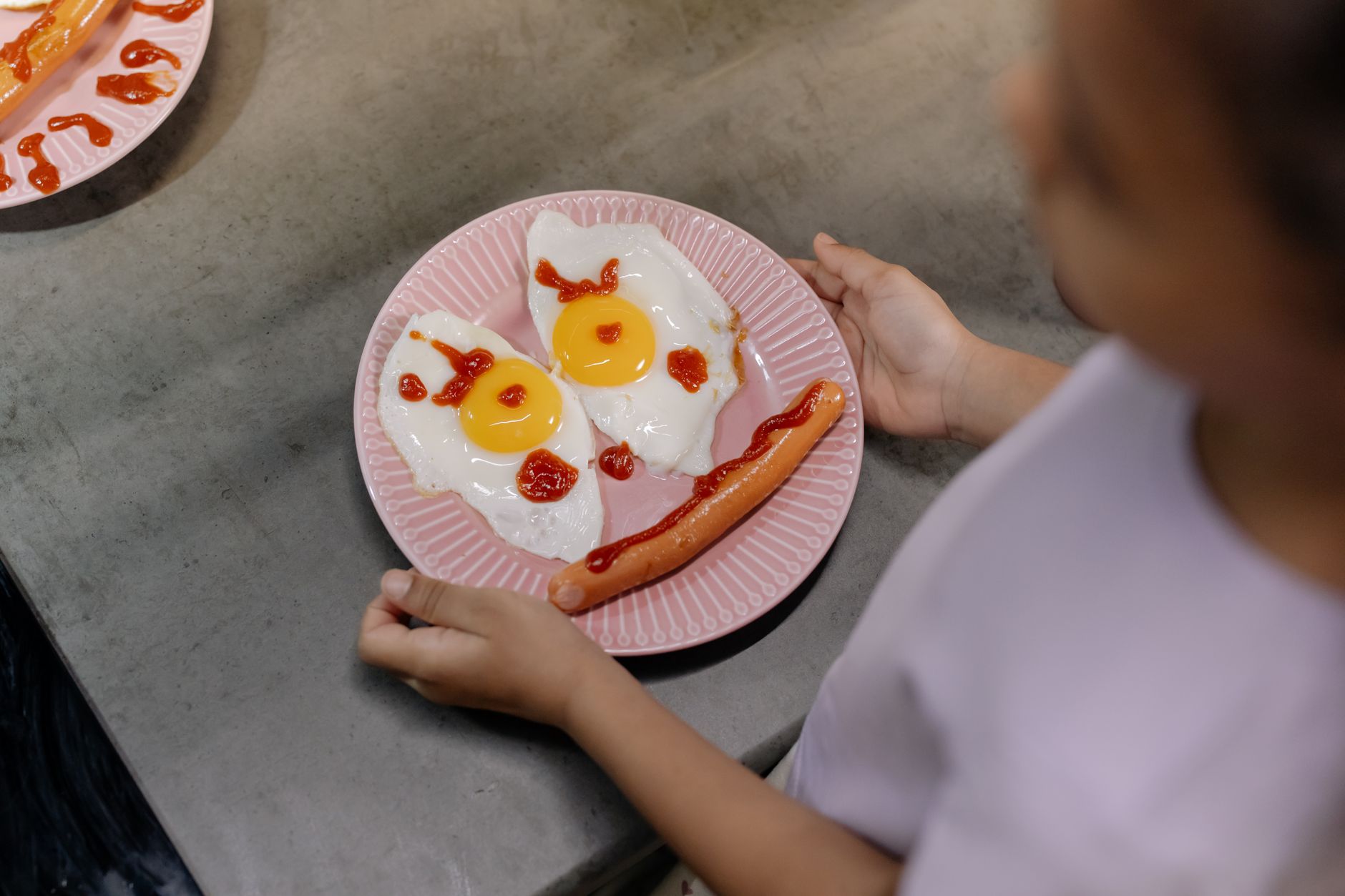 Embracing the Little Joys: Food, Fun, and Friends Playful breakfast setup featuring sunny-side-up eggs and sausage arranged like a smiling face on a pink plate.