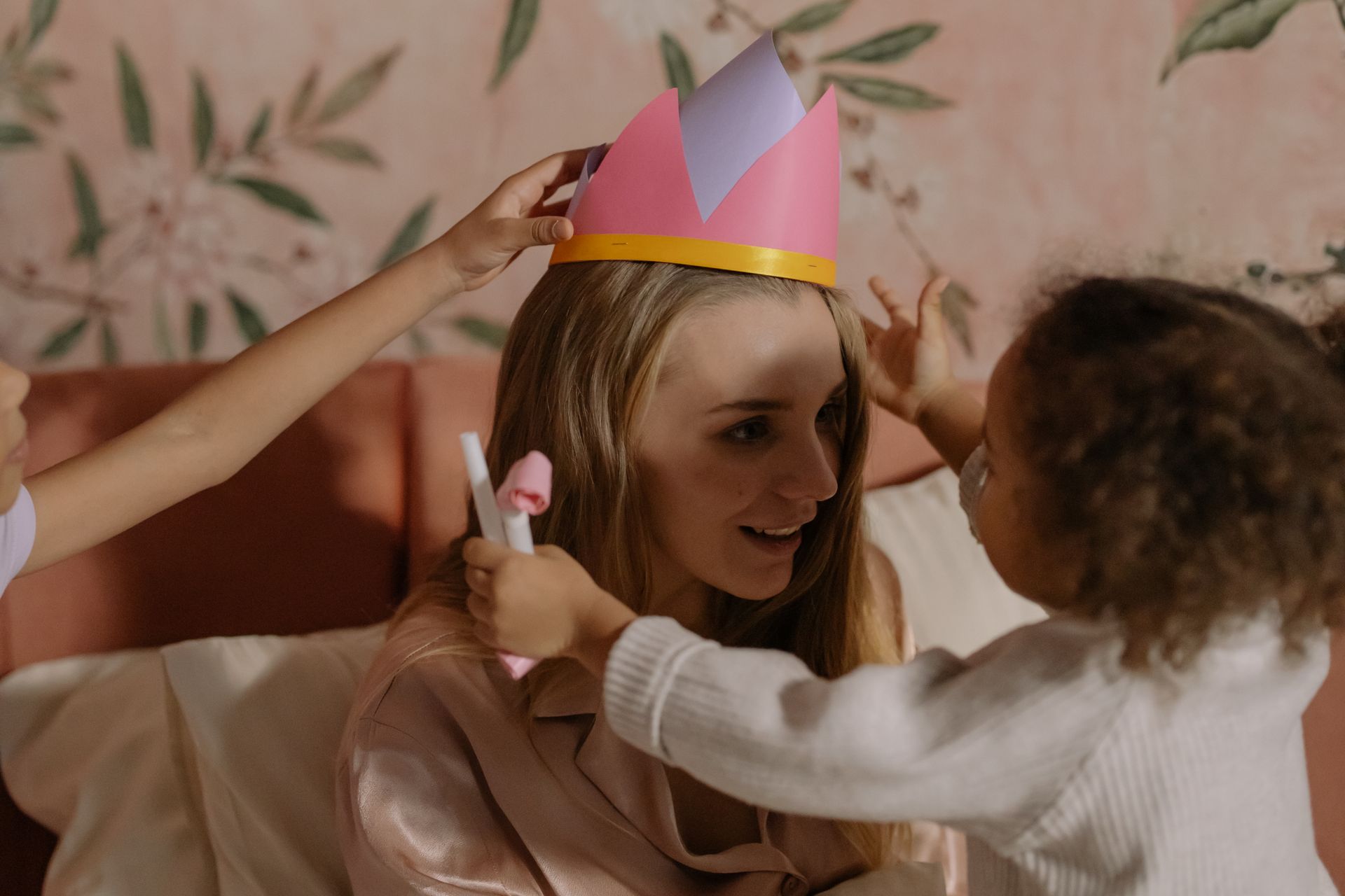 Mother and daughters share a joyful moment as they play with a paper crown indoors.
