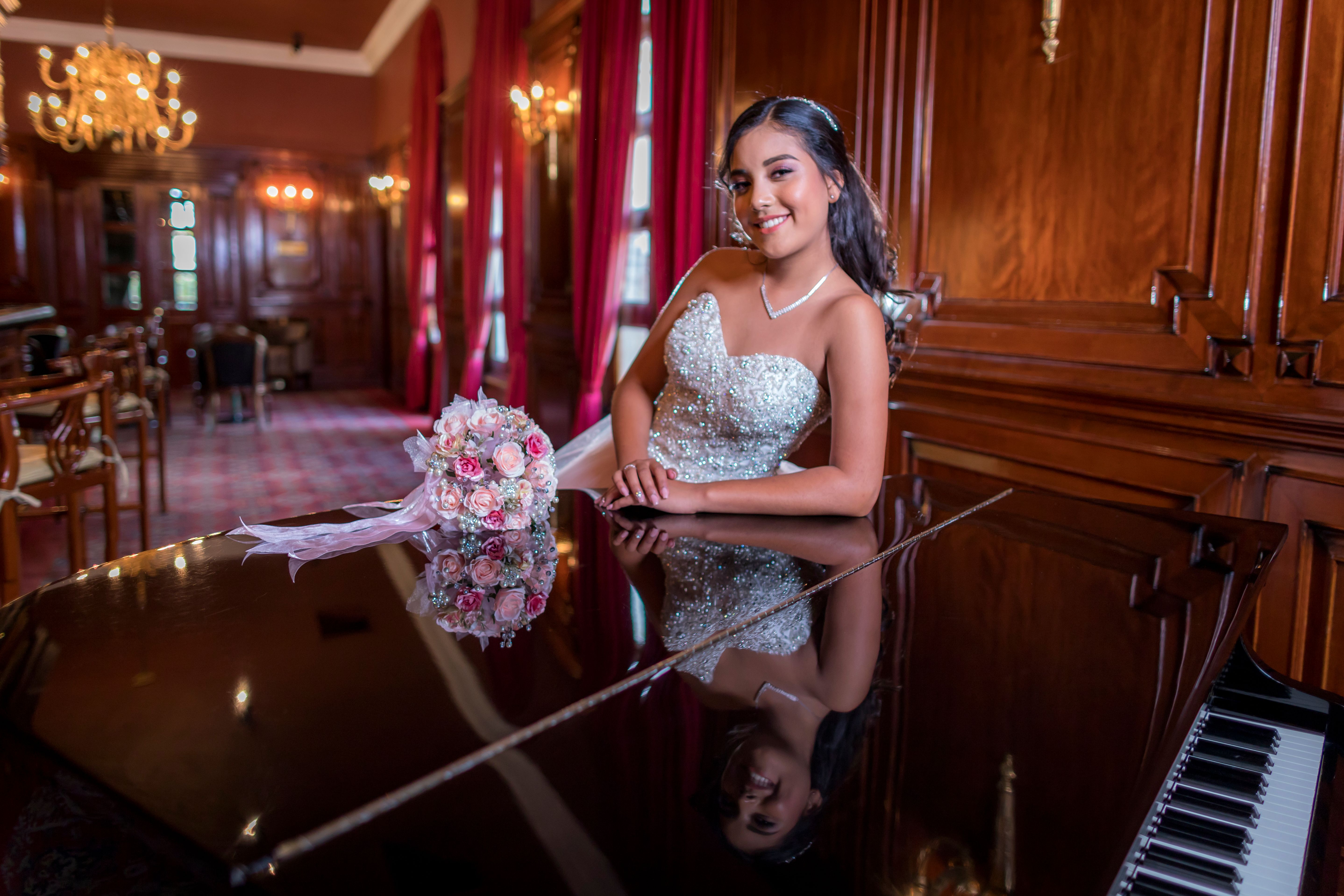 Smiling woman in elegant gown with bouquet, posing by a grand piano in a luxurious room.