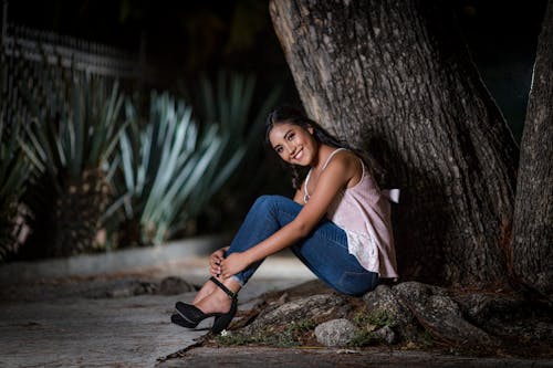 A Woman Spaghetti Strap Shirt and Blue Jeans Sitting Beside a Tree