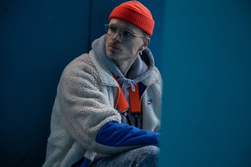 Free Serious young informal male millennial in fashionable outfit and eyeglasses sitting on haunches near blue wall and looking away thoughtfully Stock Photo