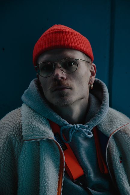Calm young unshaven guy with earring in trendy warm clothes and eyeglasses standing near blue wall and looking at camera