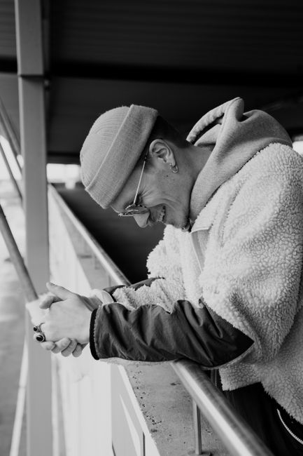 Side view of black and white positive young guy in trendy outfit and sunglasses leaning on metal railing of aged building and smiling