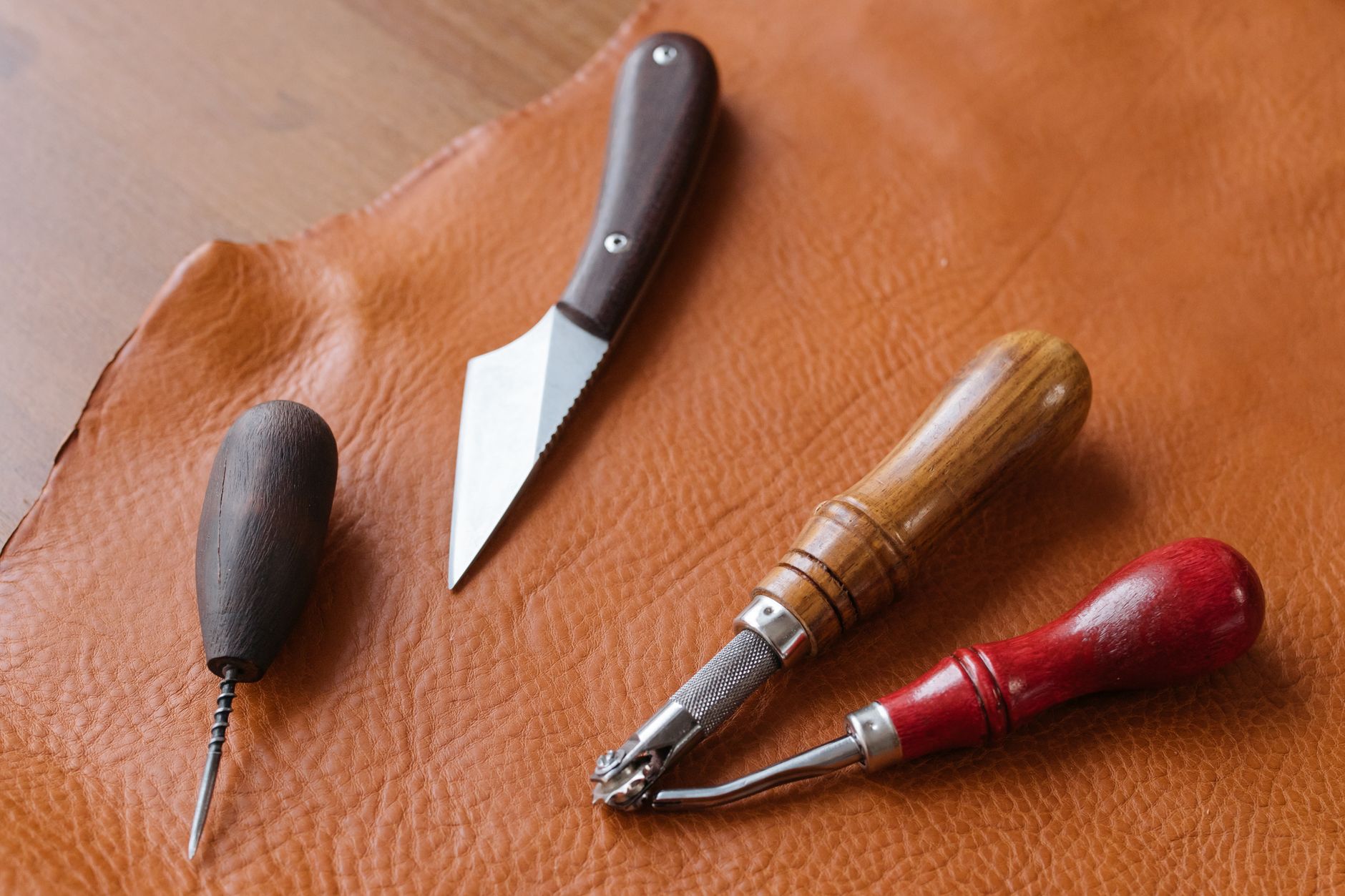 Close-up of leather crafting tools including awl, knife, and groover on a brown leather sheet.