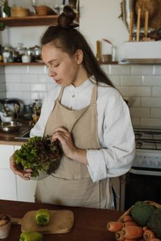Young woman in apron holding lettuce in a home kitchen, surrounded by fresh vegetables.
