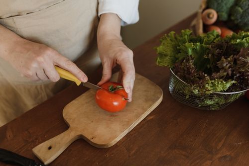 Free A person slicing a ripe tomato on a wooden board with fresh greens nearby in a kitchen setting. Stock Photo