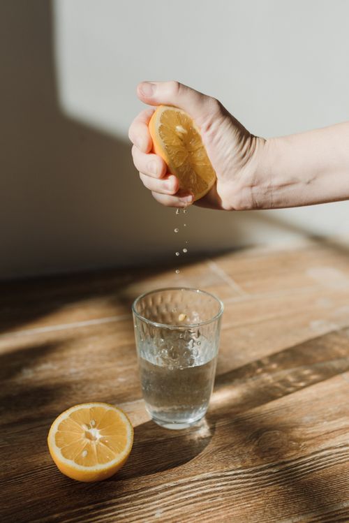 Free Hand squeezing a fresh lemon over a glass of water on a wooden surface, highlighting freshness. Stock Photo