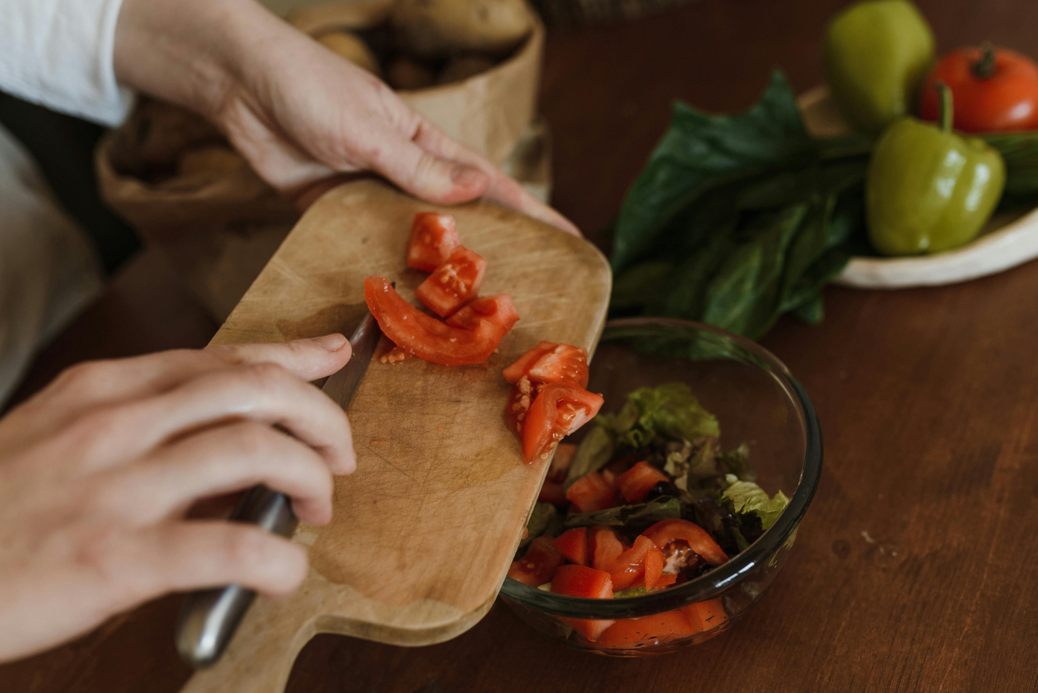 Dietary Approaches to Support Liver and Heart Health: Close-up of hands chopping tomatoes on a wooden board for a healthy salad.